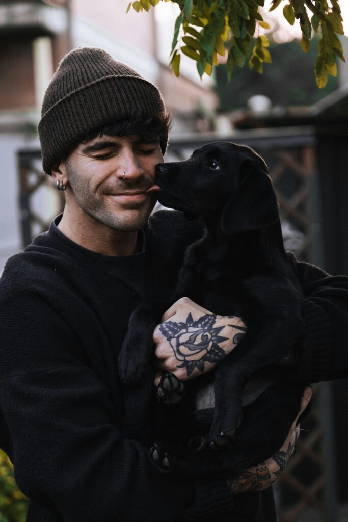 pexels-photo-30155287-30155287 Smiling man with tattoos holding a playful black Labrador puppy outdoors.